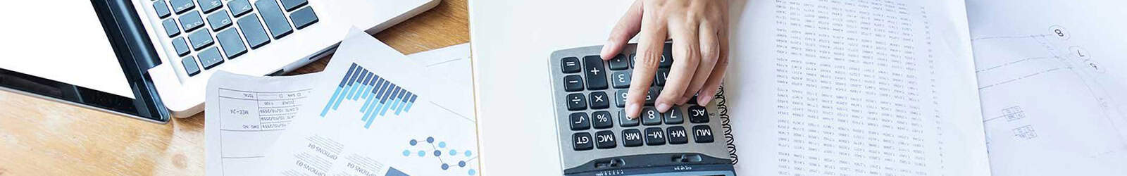 Hand typing on a calculator, surrounded by various work materials such as notebooks and a laptop