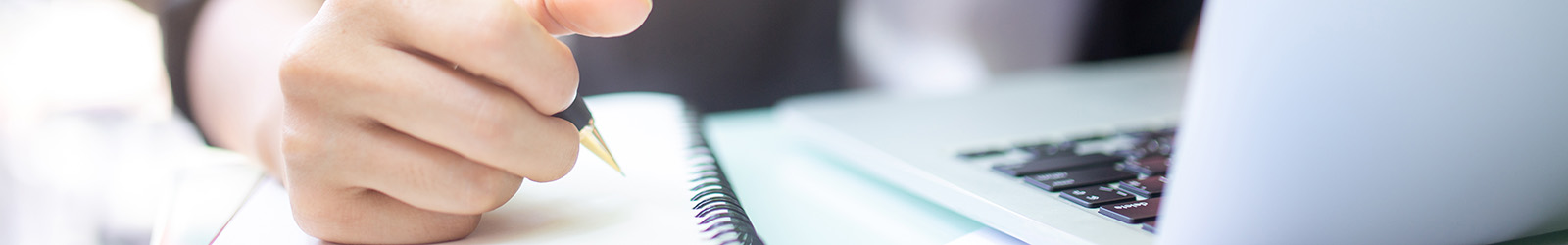 Close-up of a person's hand writing in a notebook with a pen, while a laptop is open beside them.