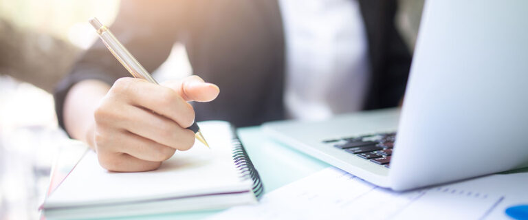 Close-up of a person's hand writing in a notebook with a pen, while a laptop is open beside them.